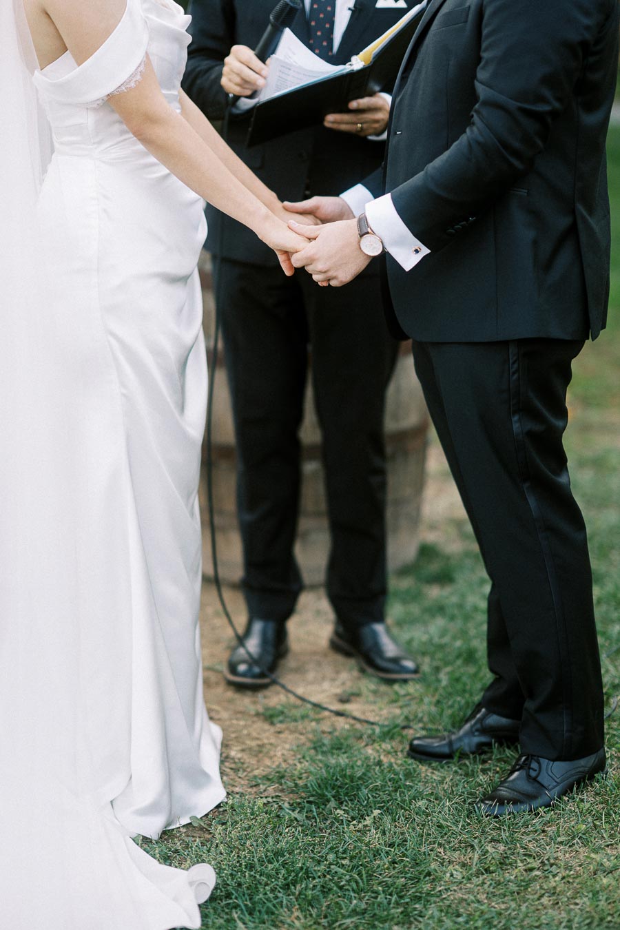 A bride and groom holding hands during their outdoor wedding ceremony, attended by an officiant, with greenery and a wine barrel in the background. The bride wears a white gown, and the groom is dressed in a black suit.