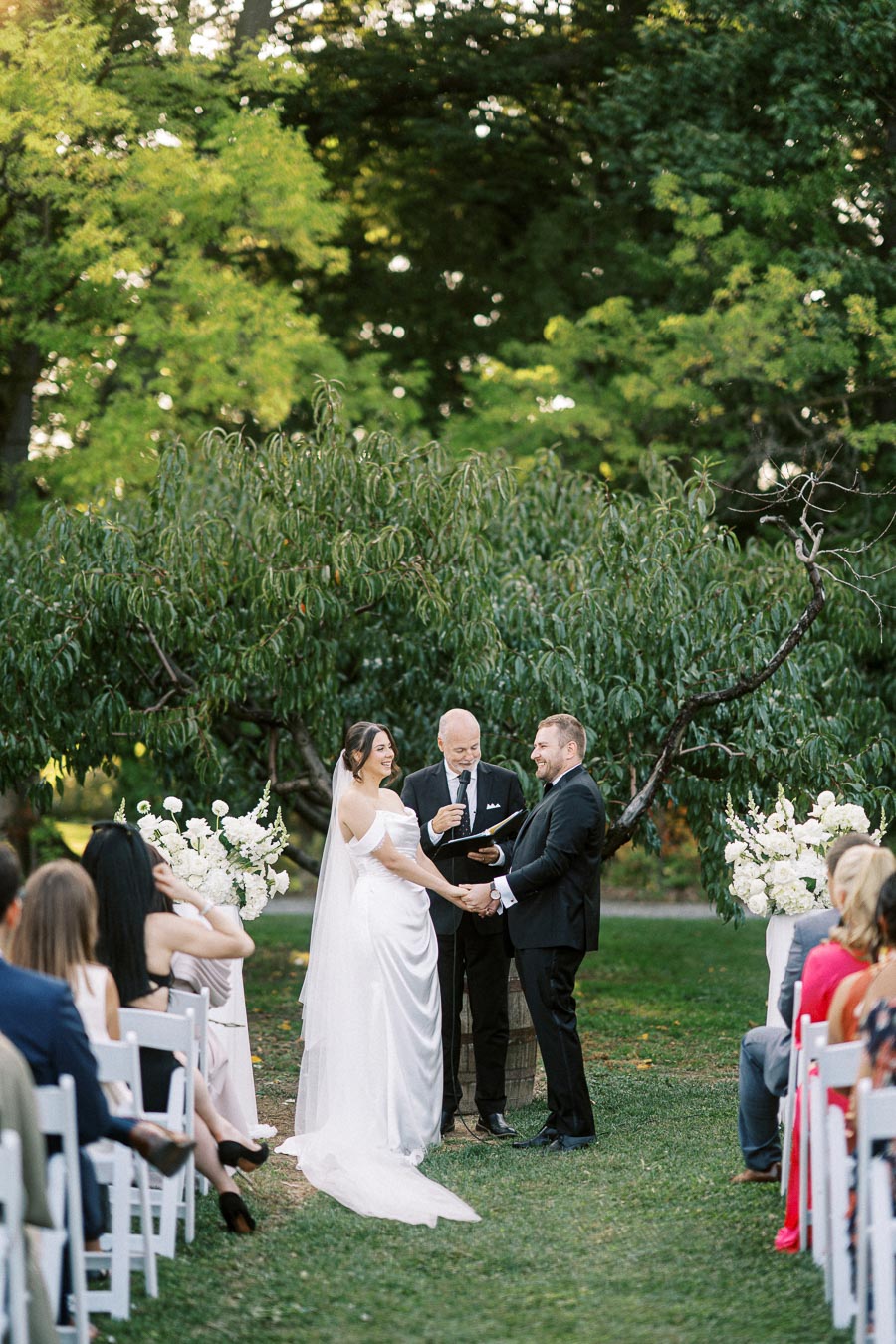 Outdoor wedding ceremony with bride and groom holding hands under a lush green arbor, surrounded by guests seated on white chairs, and officiant reading vows, set against a backdrop of vibrant trees and floral arrangements.
