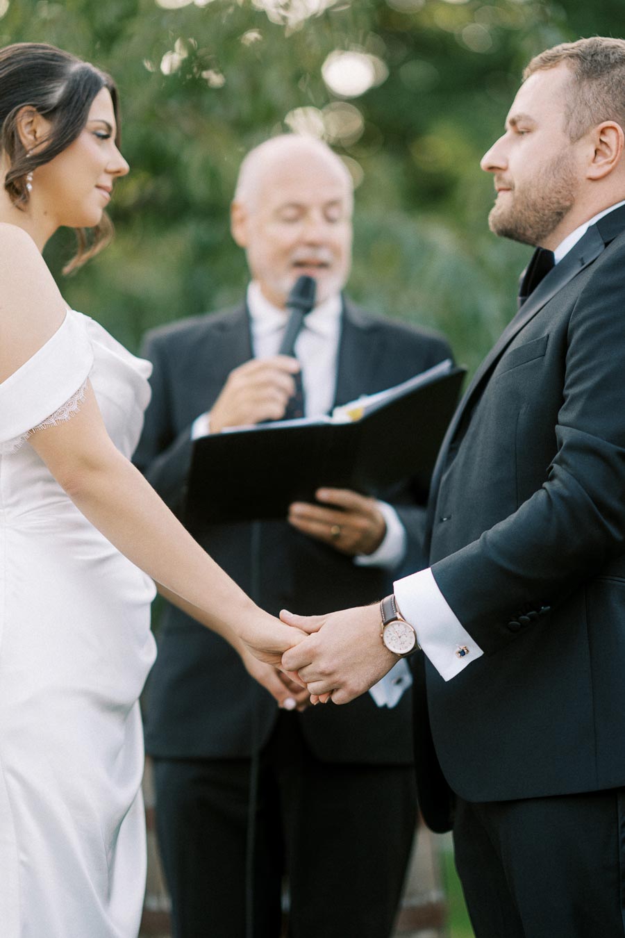 A couple holding hands during a wedding ceremony, with an officiant reading from a book in the background. The bride is wearing a white dress, and the groom is in a black suit, set against a natural outdoor backdrop.