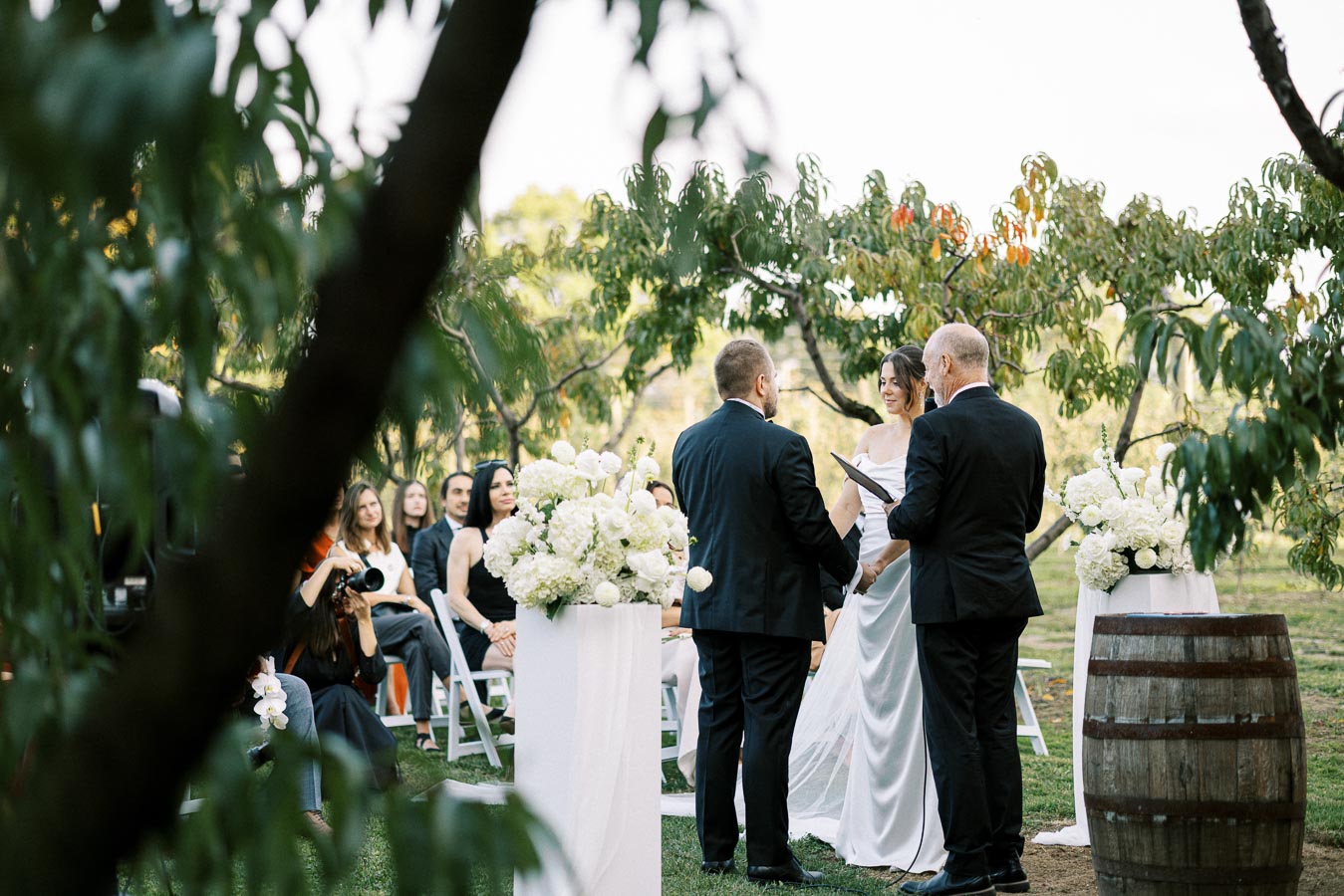 Outdoor wedding ceremony with a bride and groom exchanging vows, surrounded by guests and floral arrangements, set in a lush garden environment.