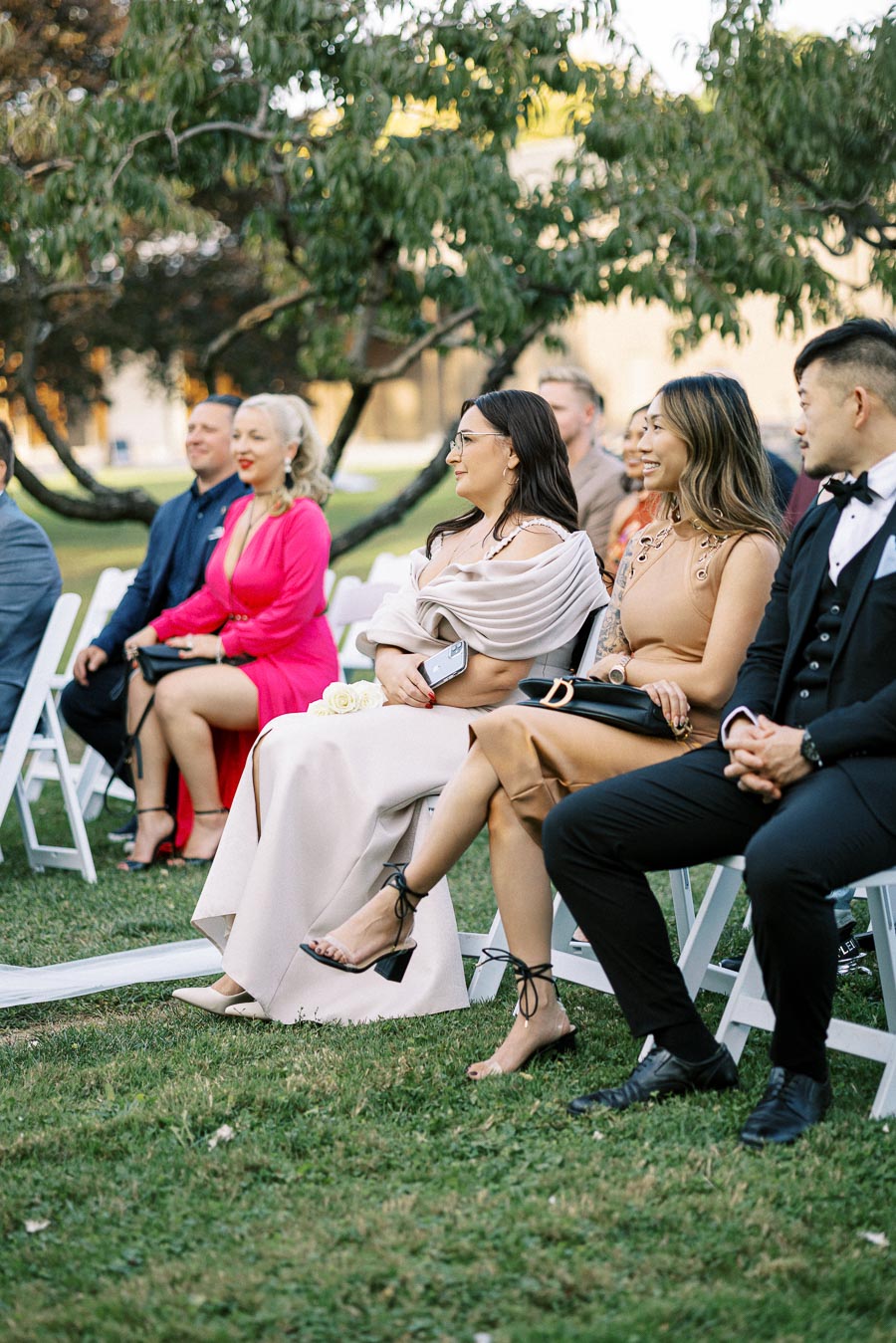 Guests seated outdoors at a wedding ceremony, dressed in formal attire, including elegant dresses and suits, with greenery in the background.