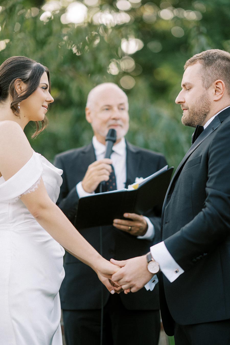 Wedding ceremony with bride and groom holding hands, officiant in background with microphone, outdoor setting.