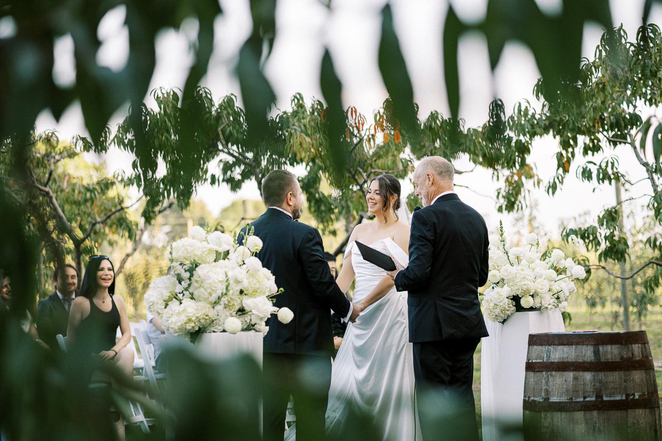 A bride and groom hold hands during an outdoor wedding ceremony, surrounded by lush greenery and elegant white floral arrangements, with the officiant standing beside them.
