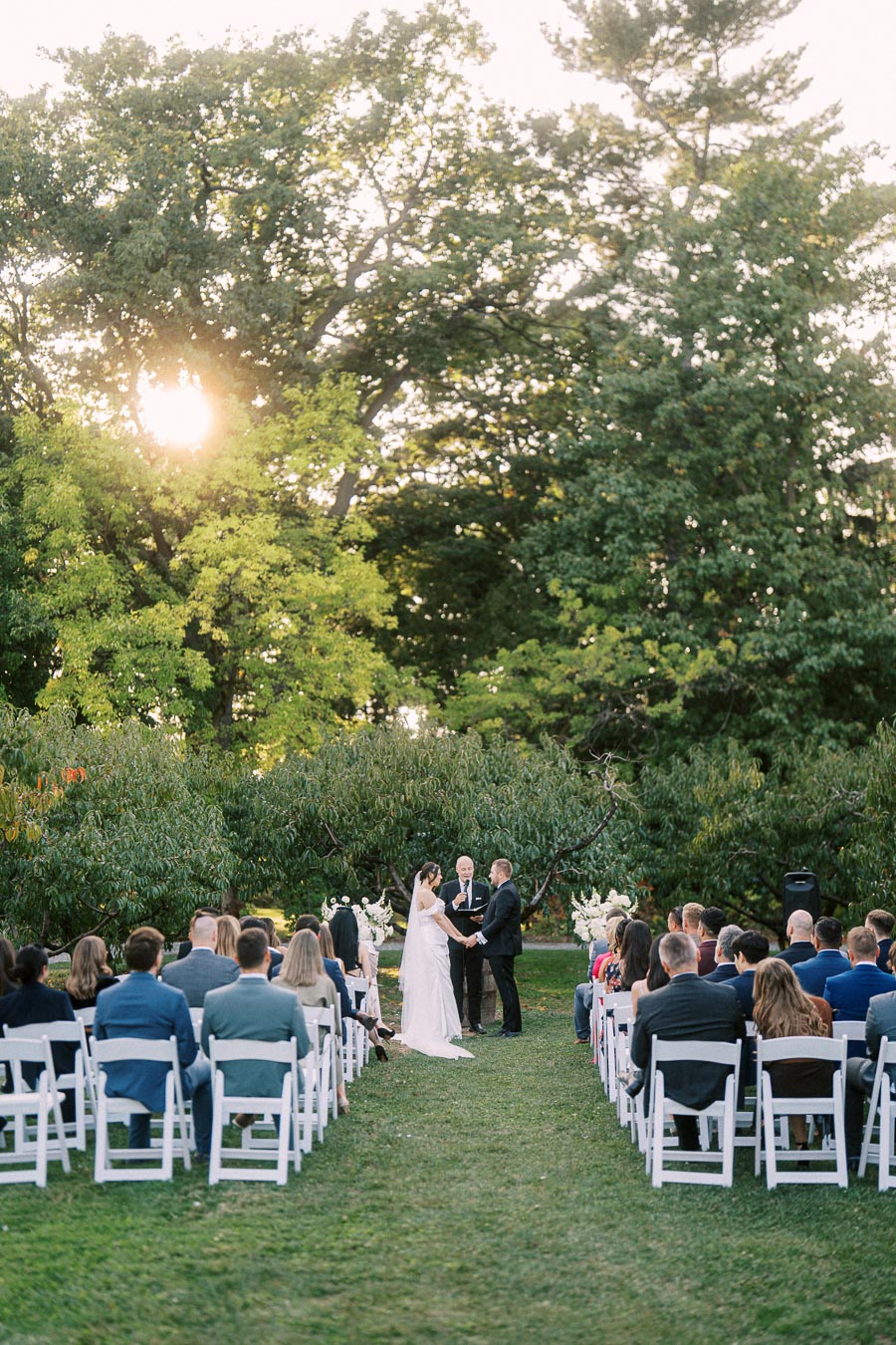 Outdoor wedding ceremony in a lush garden setting, featuring a couple exchanging vows under the warm glow of the setting sun, surrounded by guests seated in white chairs.