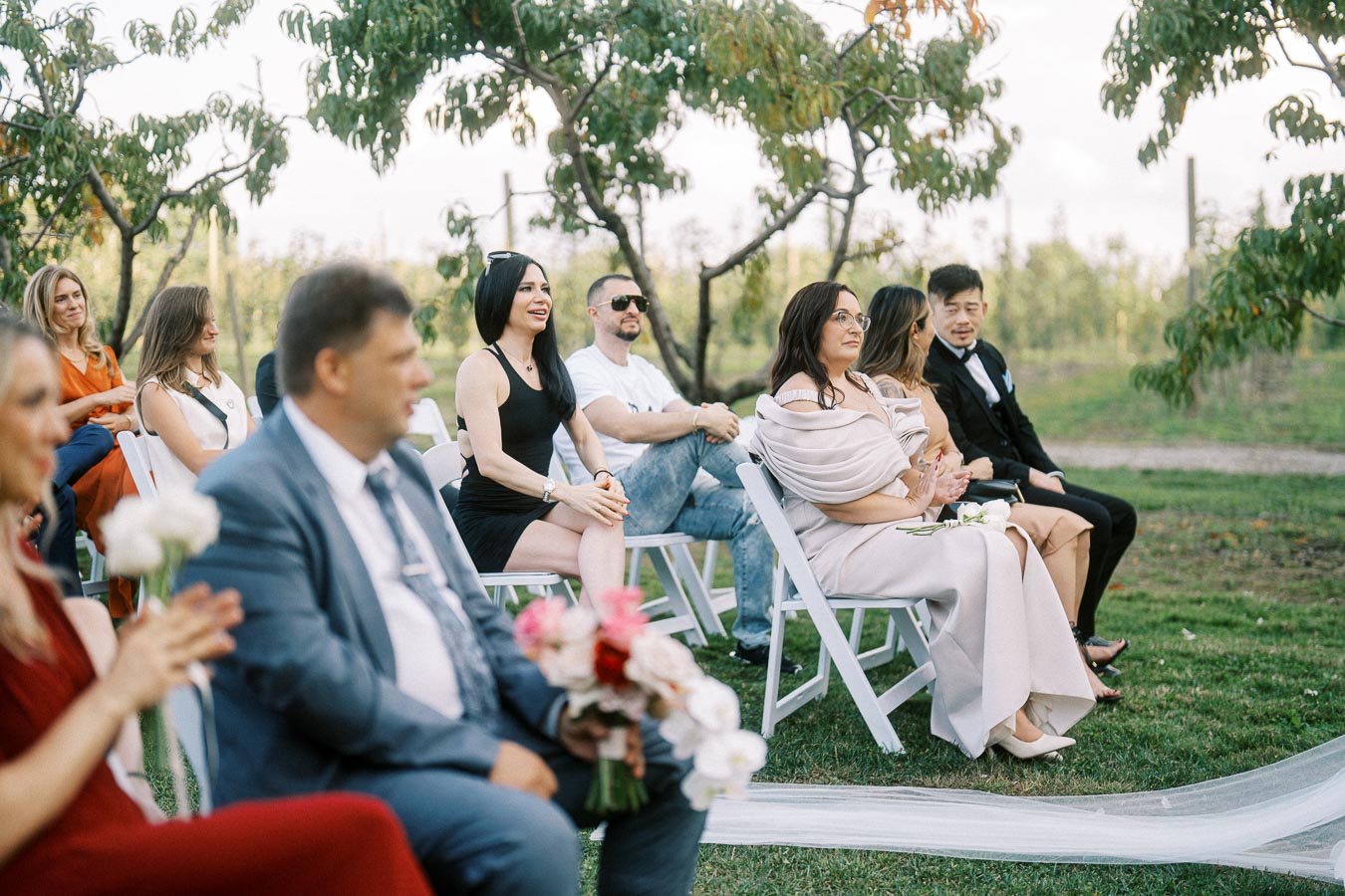 A group of elegantly dressed guests sits outdoors in a green, tree-filled setting, attentively watching an event, possibly a wedding ceremony, on a sunny day.