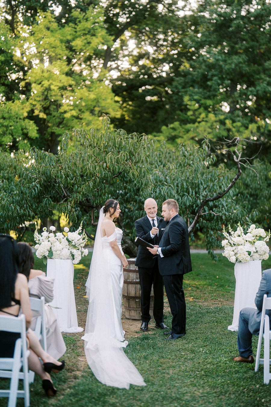 Outdoor wedding ceremony with bride in elegant white gown and groom in black suit exchanging vows. The setting features lush greenery, white floral arrangements on pedestals, and a serene natural backdrop. The officiant stands between the couple, holding a microphone and a book. Guests are seated on white chairs, attentively watching the ceremony.