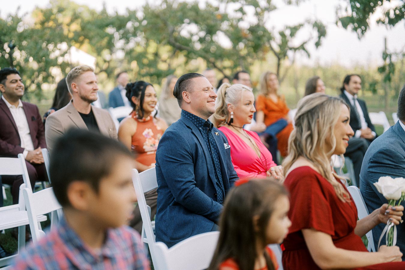 Outdoor wedding ceremony with seated guests dressed in colorful attire, enjoying the event under a sunlit garden setting.