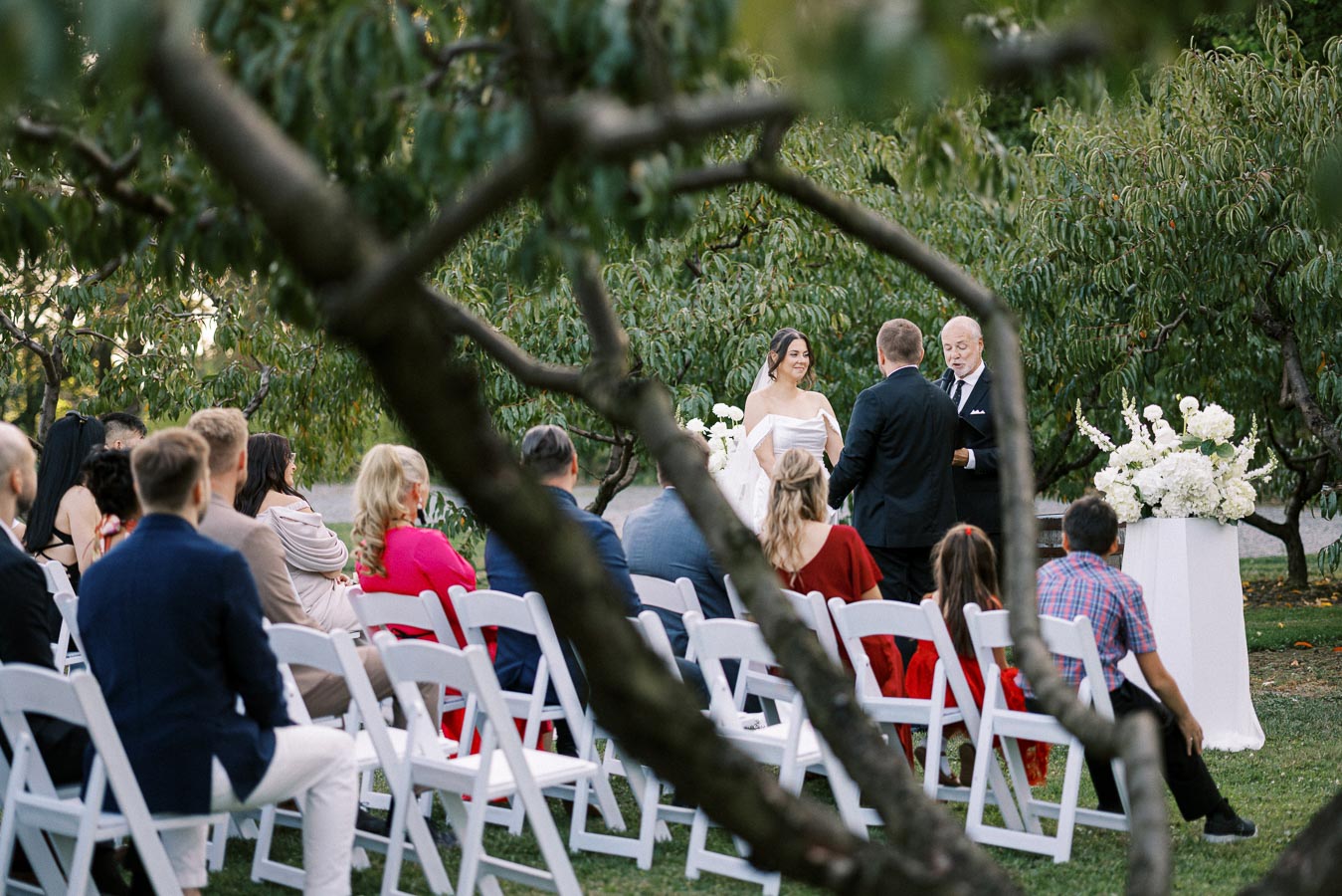 Outdoor wedding ceremony with bride and groom exchanging vows in a lush garden setting, surrounded by guests seated on white chairs.