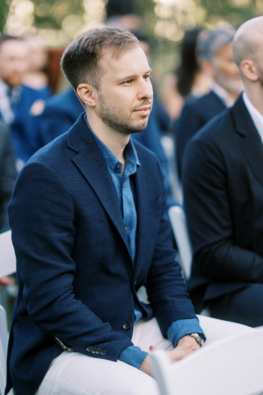 Man in a blue blazer and shirt sitting attentively during an outdoor formal event, surrounded by other attendees.