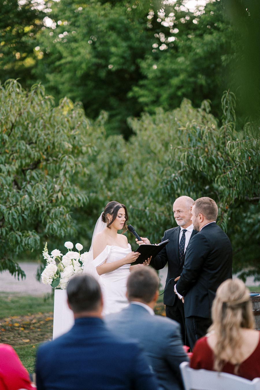 A wedding ceremony takes place outdoors, with a bride in a white gown reading her vows, accompanied by a groom in a dark suit and an officiant holding a microphone. The scene is set against a lush, green background with seated guests observing in the foreground.