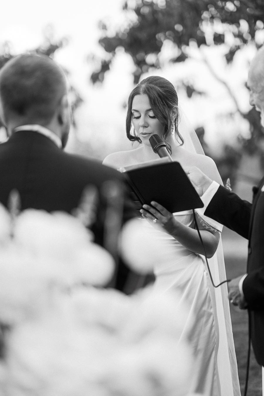 Black and white image of a bride reading her vows during an outdoor wedding ceremony, holding a microphone and standing next to the groom and officiant.