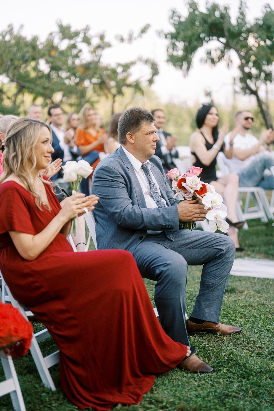 Outdoor wedding ceremony with guests sitting on white chairs. A person in a red dress and another in a grey suit holding a bouquet of flowers are attentively watching the event, surrounded by other attendees under a clear sky.