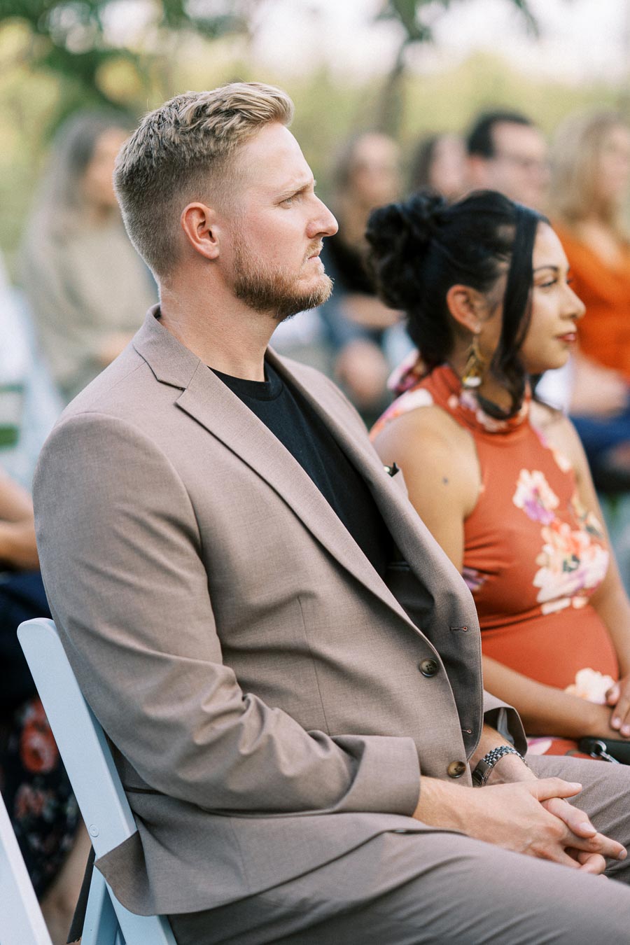 Man in a light brown suit sitting attentively during an outdoor event, accompanied by a woman in a floral dress, with blurred attendees in the background.