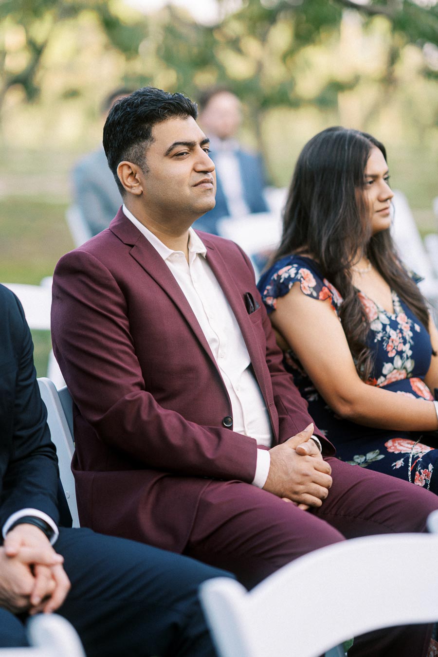Man in burgundy suit sitting at an outdoor event with blurred background