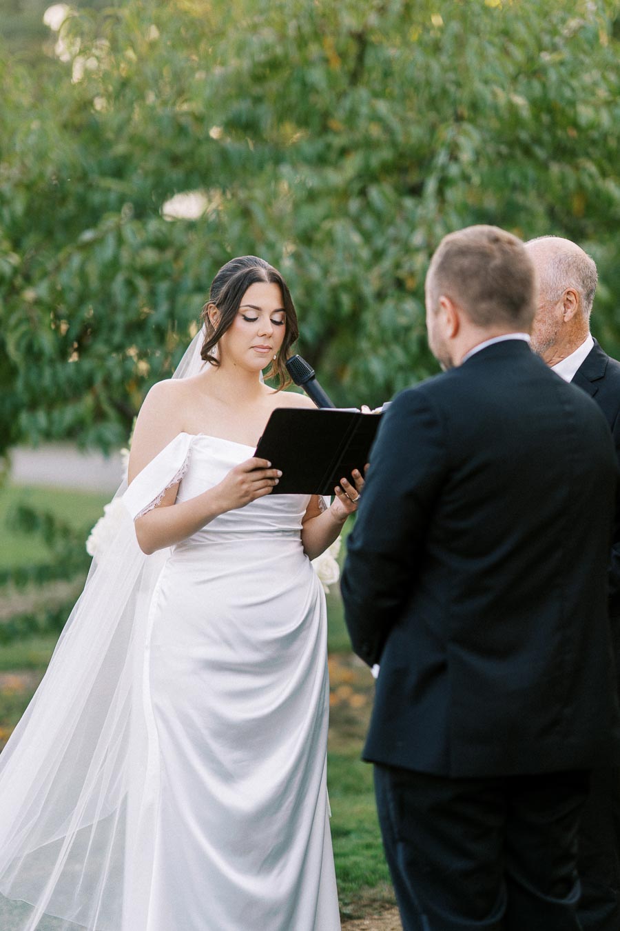 Bride in a white wedding dress reads vows from a book during an outdoor ceremony, facing her groom and officiant.