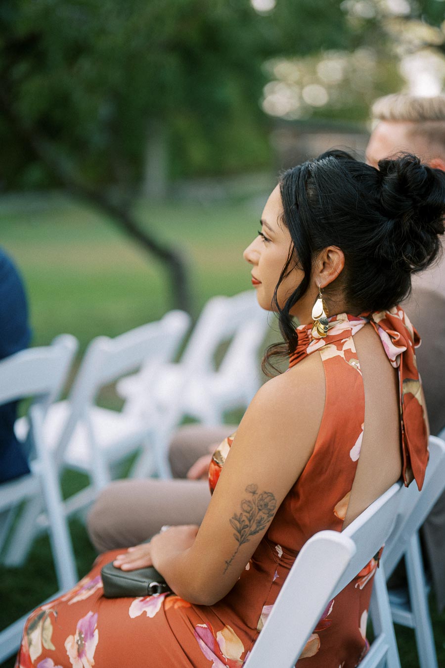 Elegant woman in a floral dress attending an outdoor event, seated on a white chair.