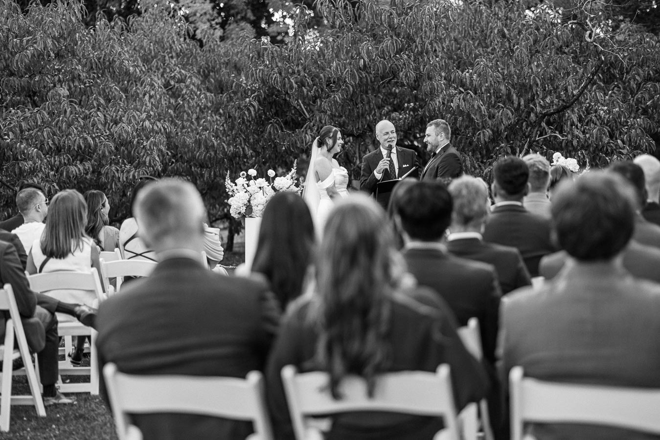 Black and white outdoor wedding ceremony with bride and groom standing in front of officiant, surrounded by seated guests in a garden setting with lush greenery.