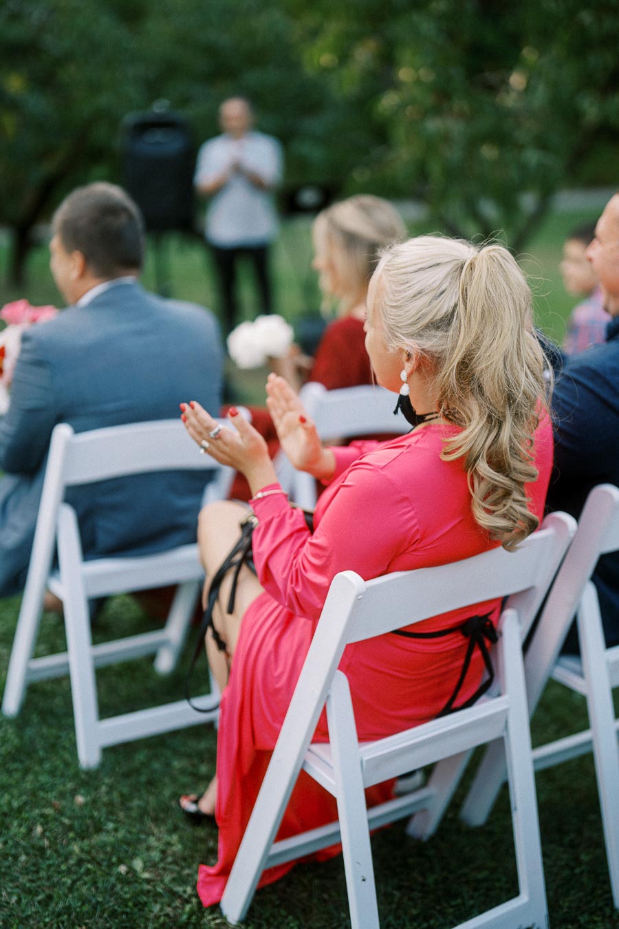 Outdoor event with attendees seated on white chairs, applauding while a speaker is visible in the background. The focus is on a woman in a pink dress with blond hair, emphasizing a celebratory and engaging atmosphere.