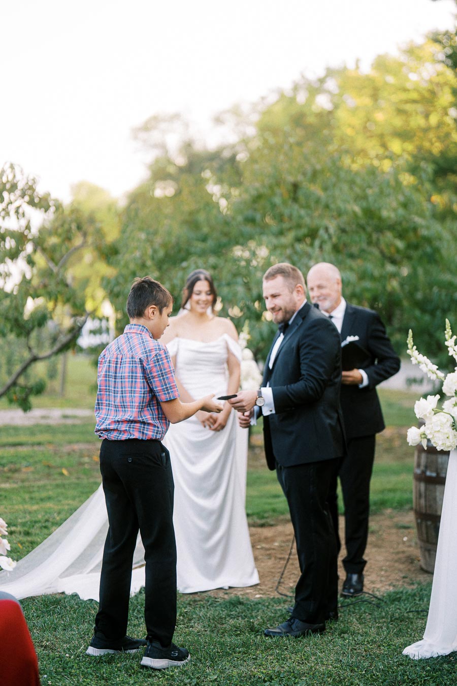 A young boy hands over a ring to a groom during an outdoor wedding ceremony, with the bride and officiant standing nearby, surrounded by greenery and flowers.