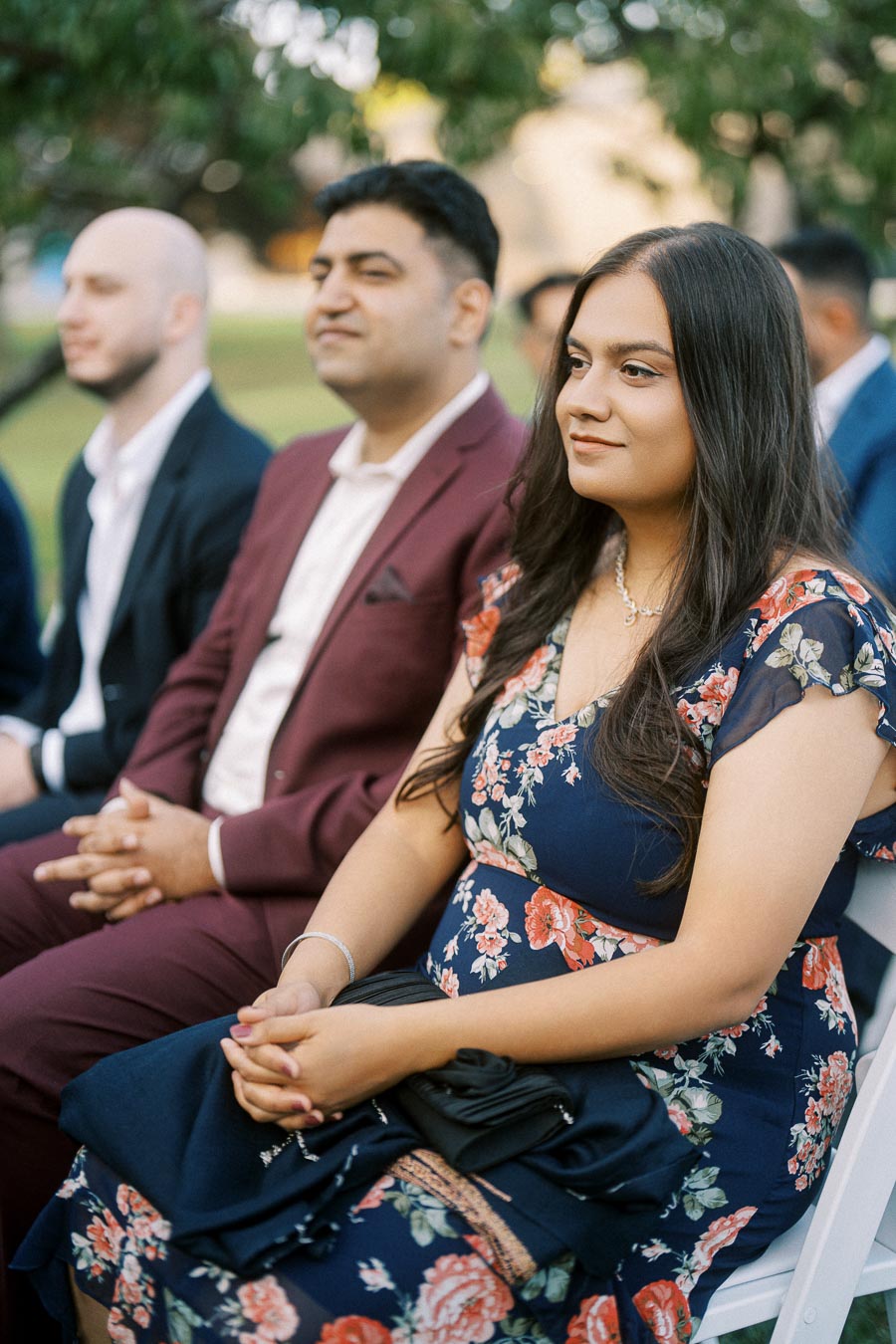 People attentively sitting at an outdoor event, with a woman in a floral dress in focus, surrounded by others in formal attire.