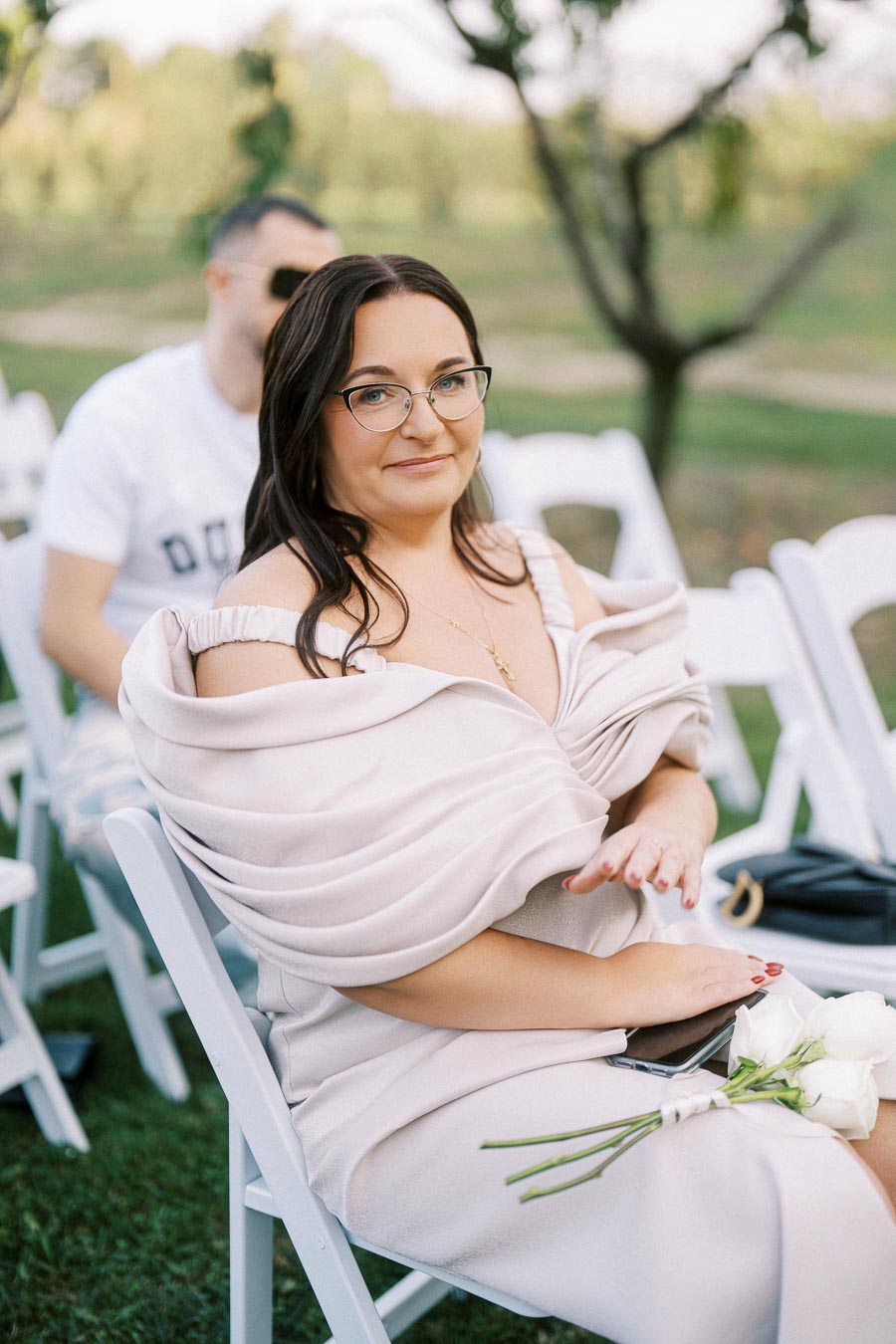 A woman in glasses wearing an elegant off-shoulder dress seated on a white chair outdoors, holding a phone and a small bouquet of white flowers.