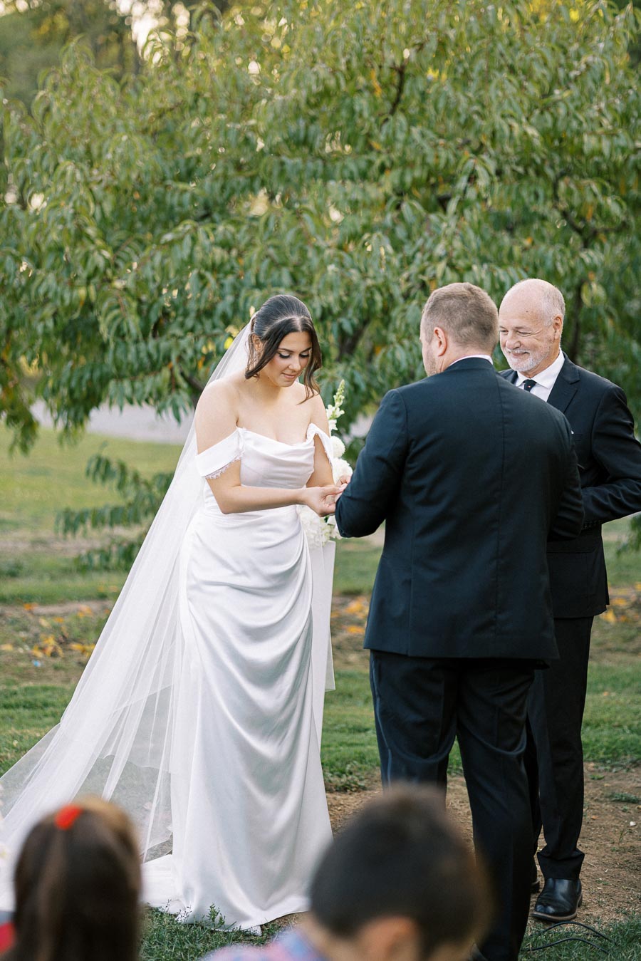 Bride and groom exchanging vows in a garden ceremony, with a lush green backdrop and a smiling officiant, during a sunny outdoor wedding.