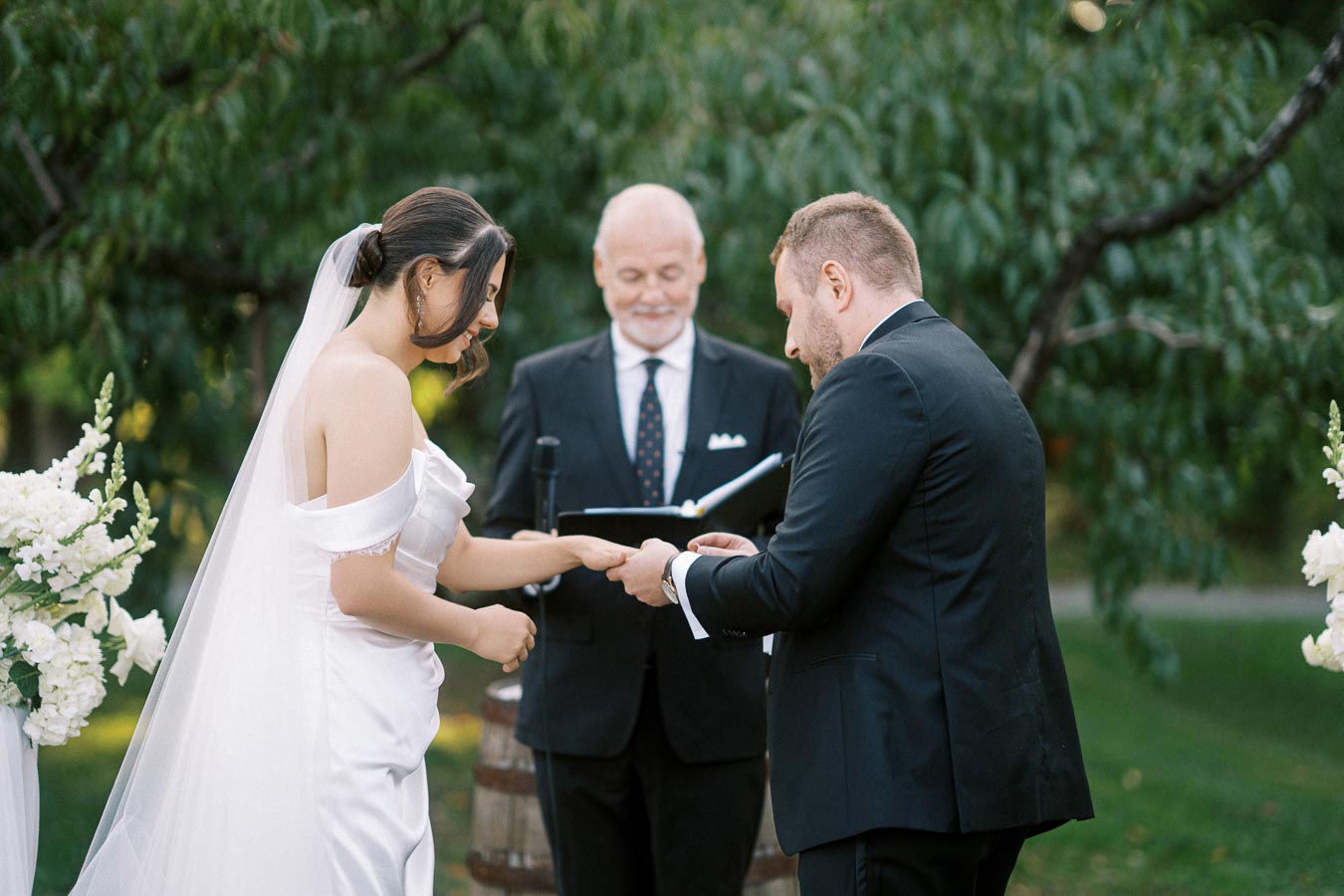 A bride and groom exchange rings during an outdoor wedding ceremony, with an officiant standing behind them, surrounded by greenery and white floral arrangements.