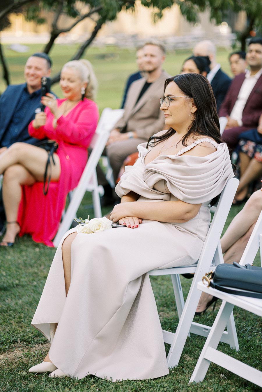 Guests seated outdoors at a wedding ceremony, with a woman in an elegant beige dress holding a white rose on her lap, surrounded by other attendees on a sunny day.