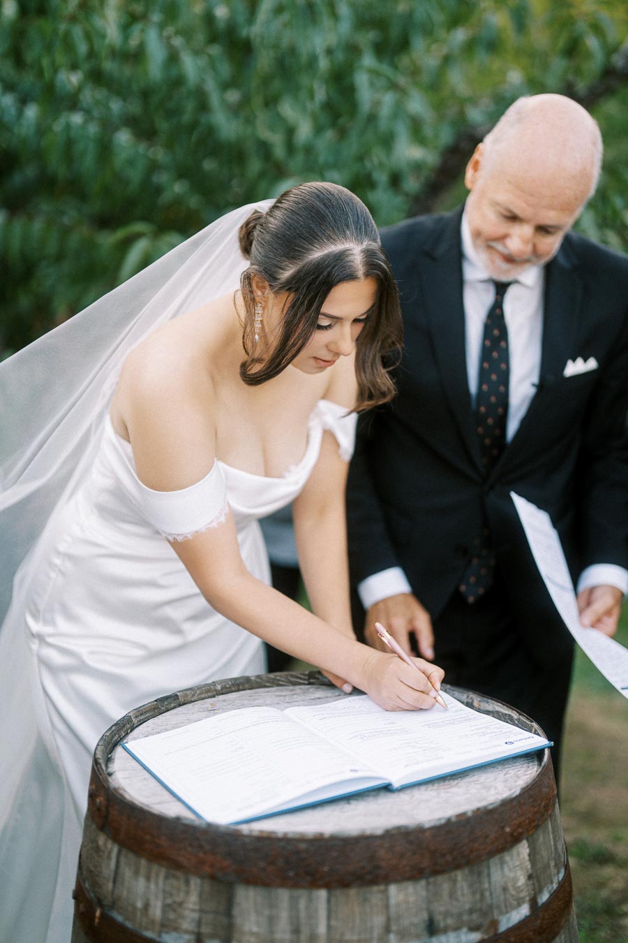 Bride in elegant white gown signs wedding document on rustic wooden barrel outdoors, accompanied by a man in a black suit.