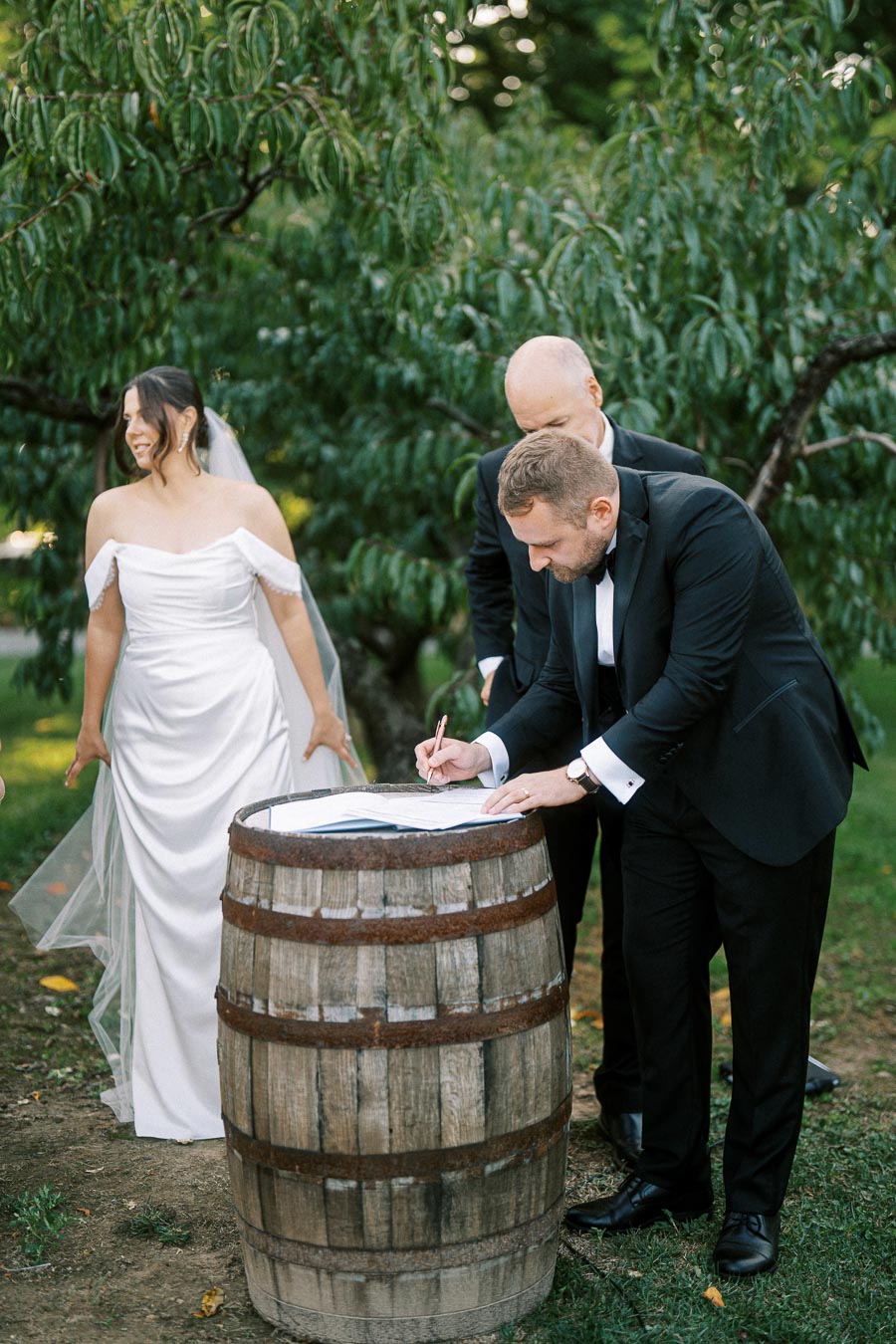 Bride and groom signing marriage certificate on a rustic wooden barrel in an outdoor garden setting.