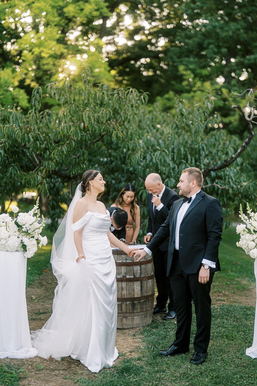 A bride in a white gown and groom in a black tuxedo holding hands during an outdoor wedding ceremony, surrounded by lush greenery and white floral arrangements.
