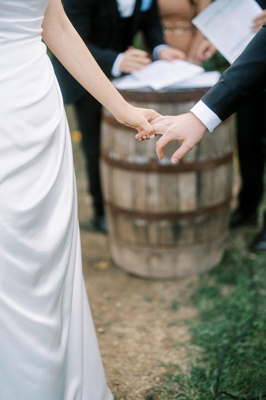 Wedding ceremony moment with bride in a white dress and groom in a black suit holding hands, with a rustic wooden barrel table in the background.