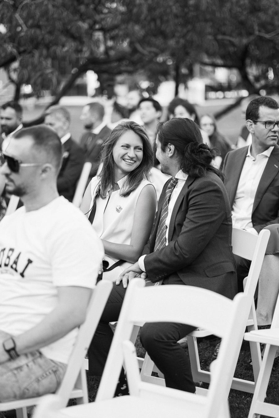Black and white photo of a woman smiling at a man during an outdoor event, seated amongst attendees on white folding chairs.