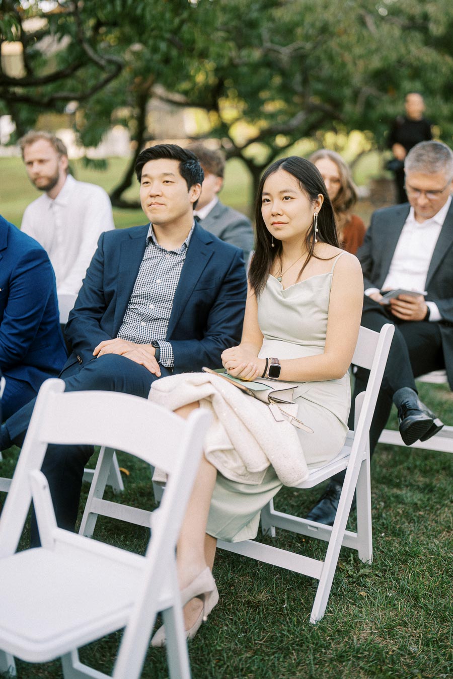 Attentive guests seated at an outdoor event, with one man and woman in formal attire, surrounded by greenery and other attendees in the background.