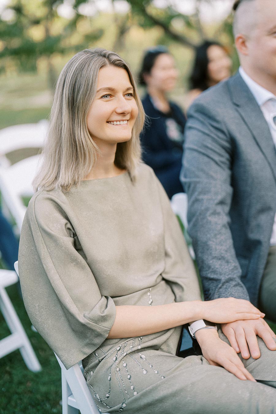 Young woman in a light green dress smiling while sitting outdoors at an event with people seated in the background.