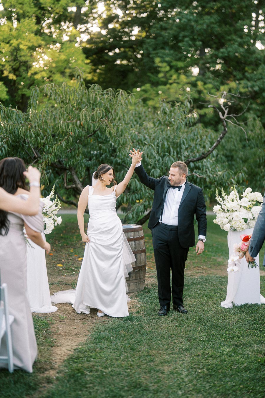 A joyful bride and groom celebrate their outdoor wedding ceremony with a high-five, surrounded by lush greenery and guests.