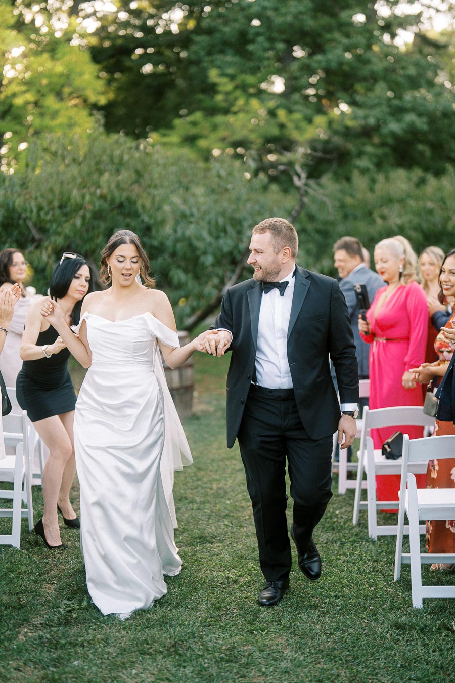 Wedding couple walking down the aisle, bride in white dress and groom in black tuxedo, surrounded by cheering guests at an outdoor ceremony.