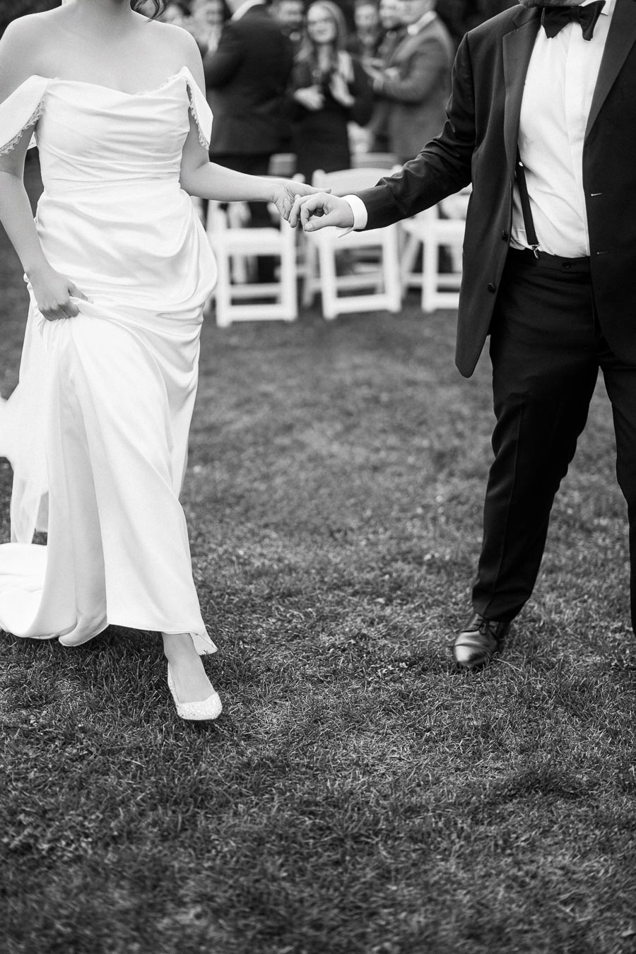 Black and white photo of a bride in a white wedding dress and a groom in a suit holding hands while walking on grass during an outdoor ceremony.