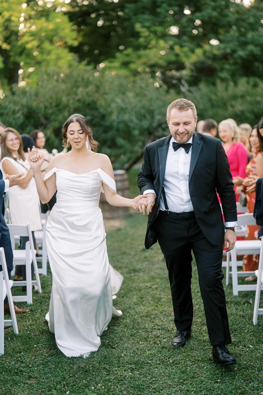A joyful couple holding hands and walking down the aisle at an outdoor wedding ceremony, surrounded by smiling guests and lush greenery. The bride is wearing an elegant off-shoulder white wedding gown, and the groom is donning a classic black tuxedo.