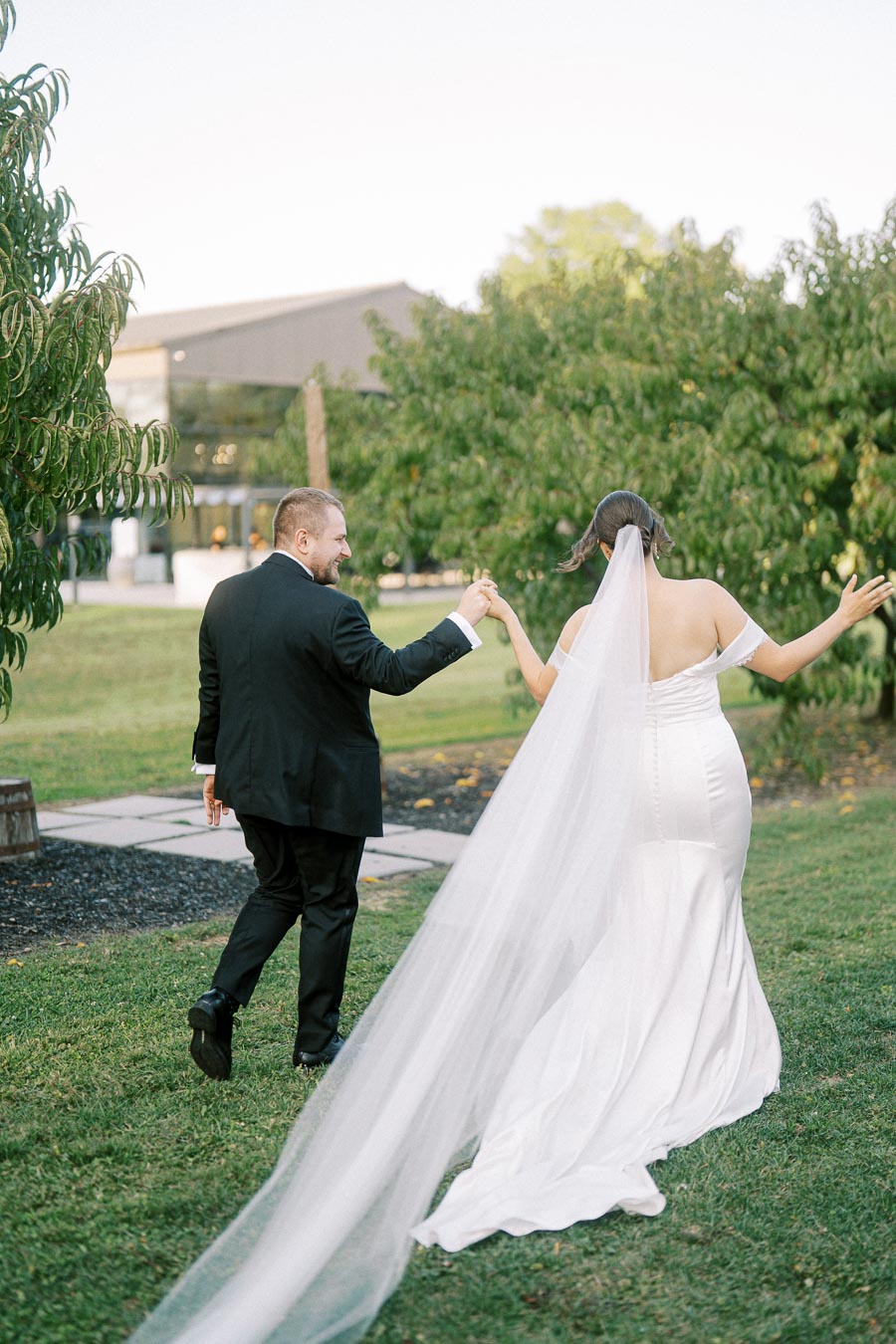 A bride and groom holding hands, walking through a lush garden in wedding attire, with the bride wearing a long white gown and veil.