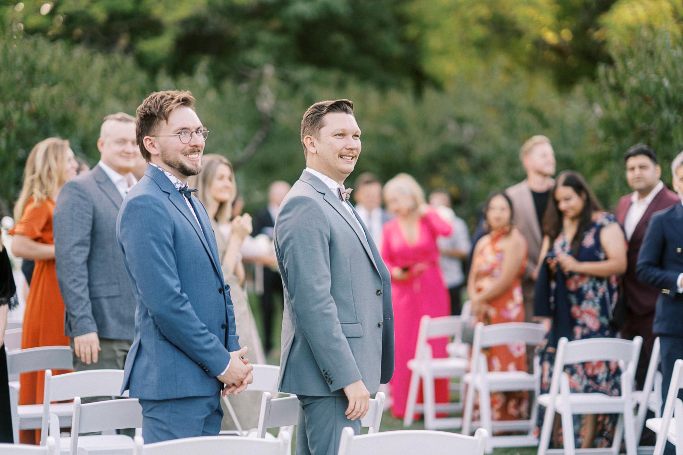 Two groomsmen in suits smiling at an outdoor wedding ceremony, surrounded by seated guests in formal attire.