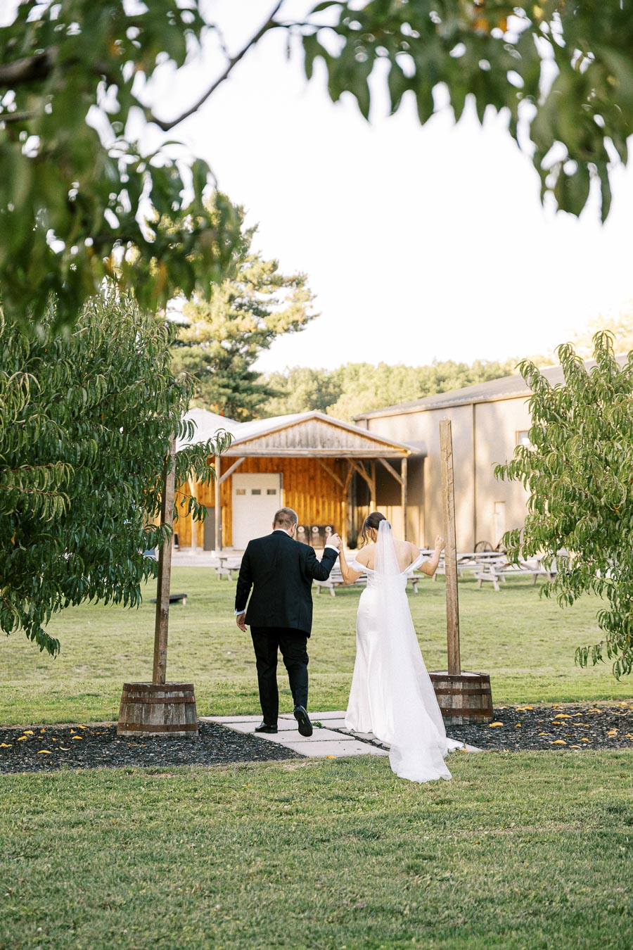 Bride and groom walking hand in hand towards a rustic barn venue, surrounded by lush greenery and trees, on a sunny wedding day.