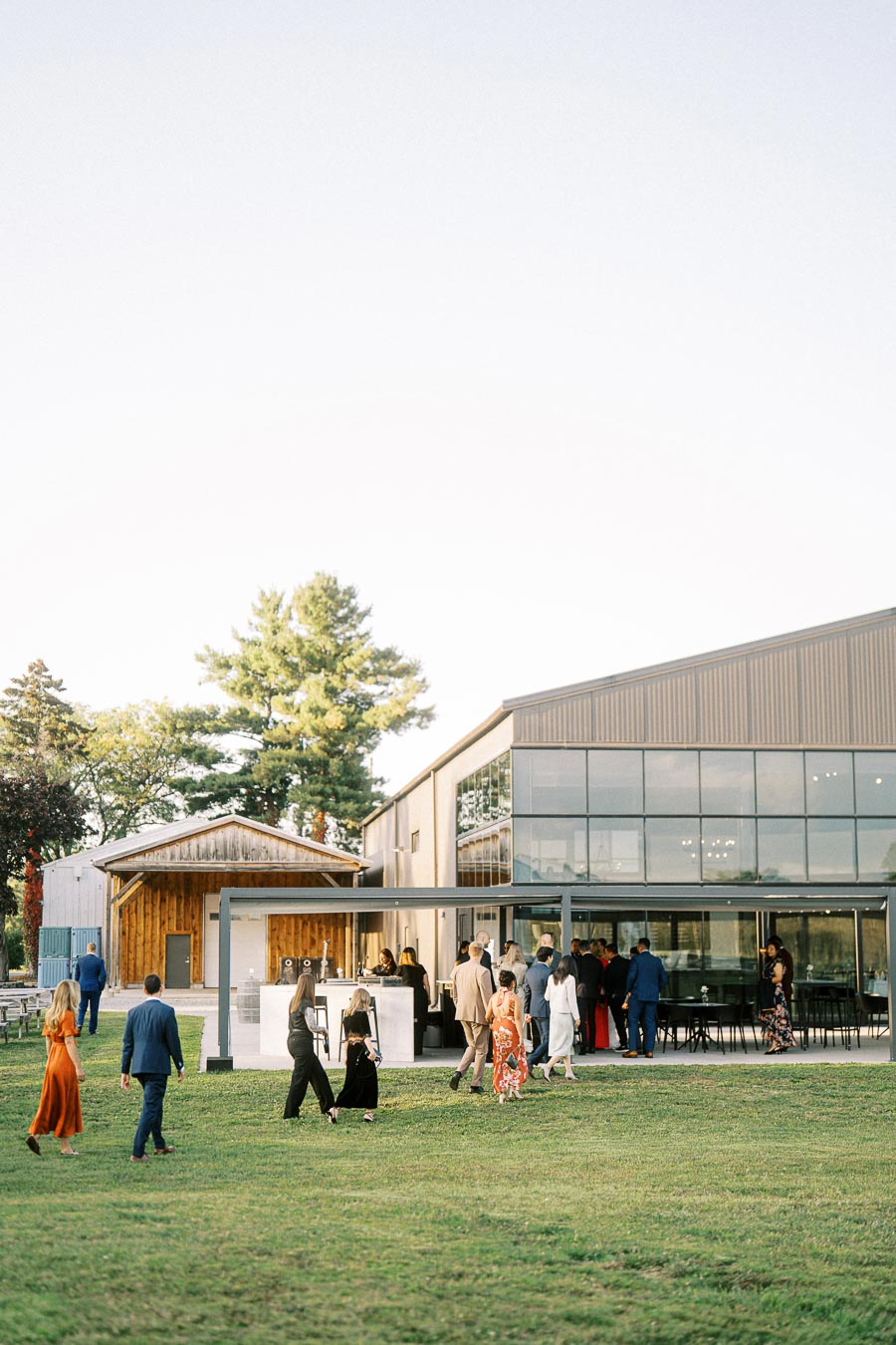 A group of people walking towards a modern glass-walled building set in a scenic outdoor area with green grass and trees under a clear sky, dressed in formal attire for what appears to be an event or gathering.