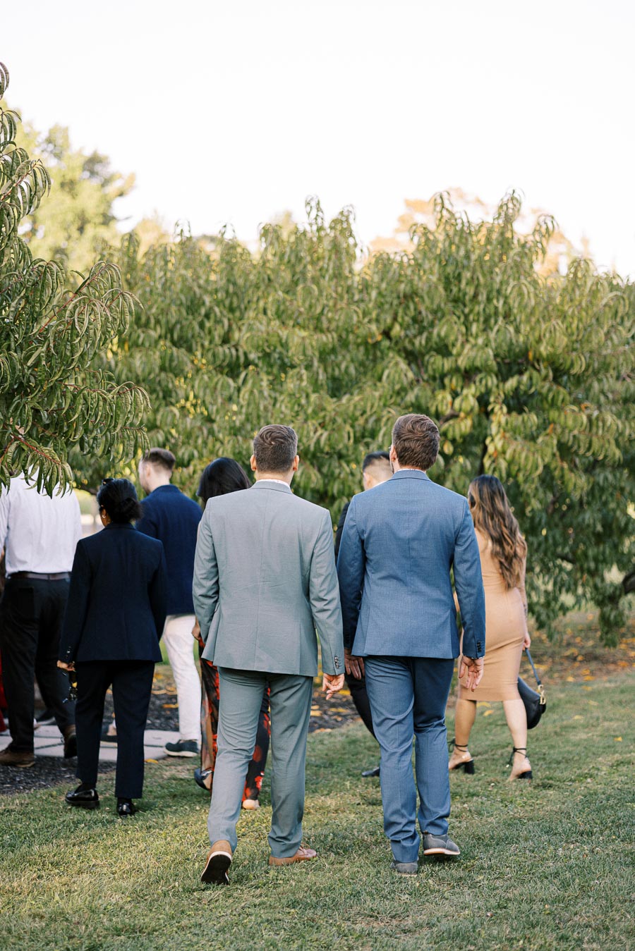 A group of people in formal attire walking through a garden setting, with lush green trees and a clear sky, creating a serene outdoor gathering atmosphere.
