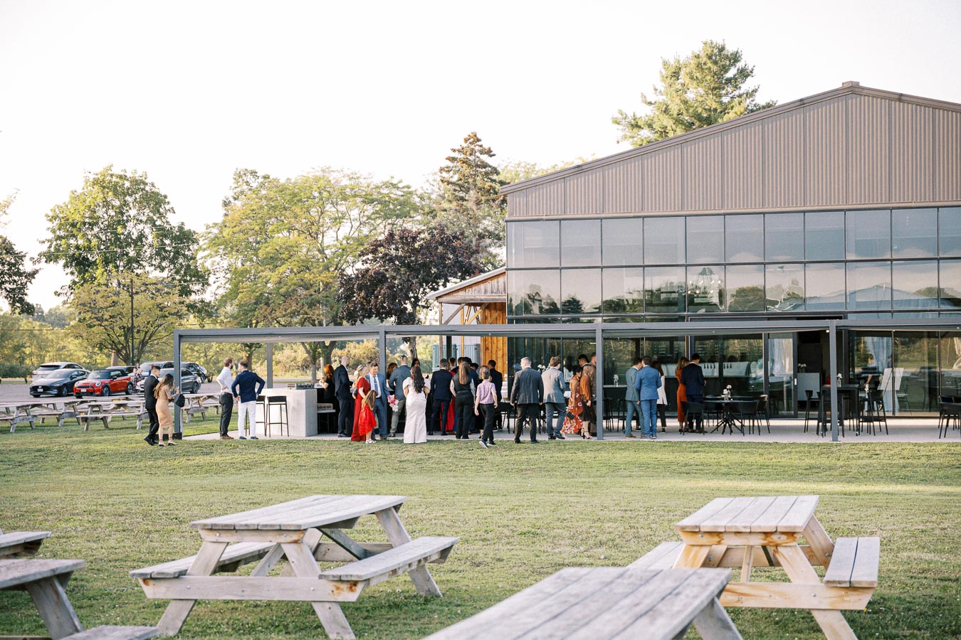 Outdoor wedding reception at a modern glass venue with guests socializing near a bar, surrounded by greenery and picnic tables.