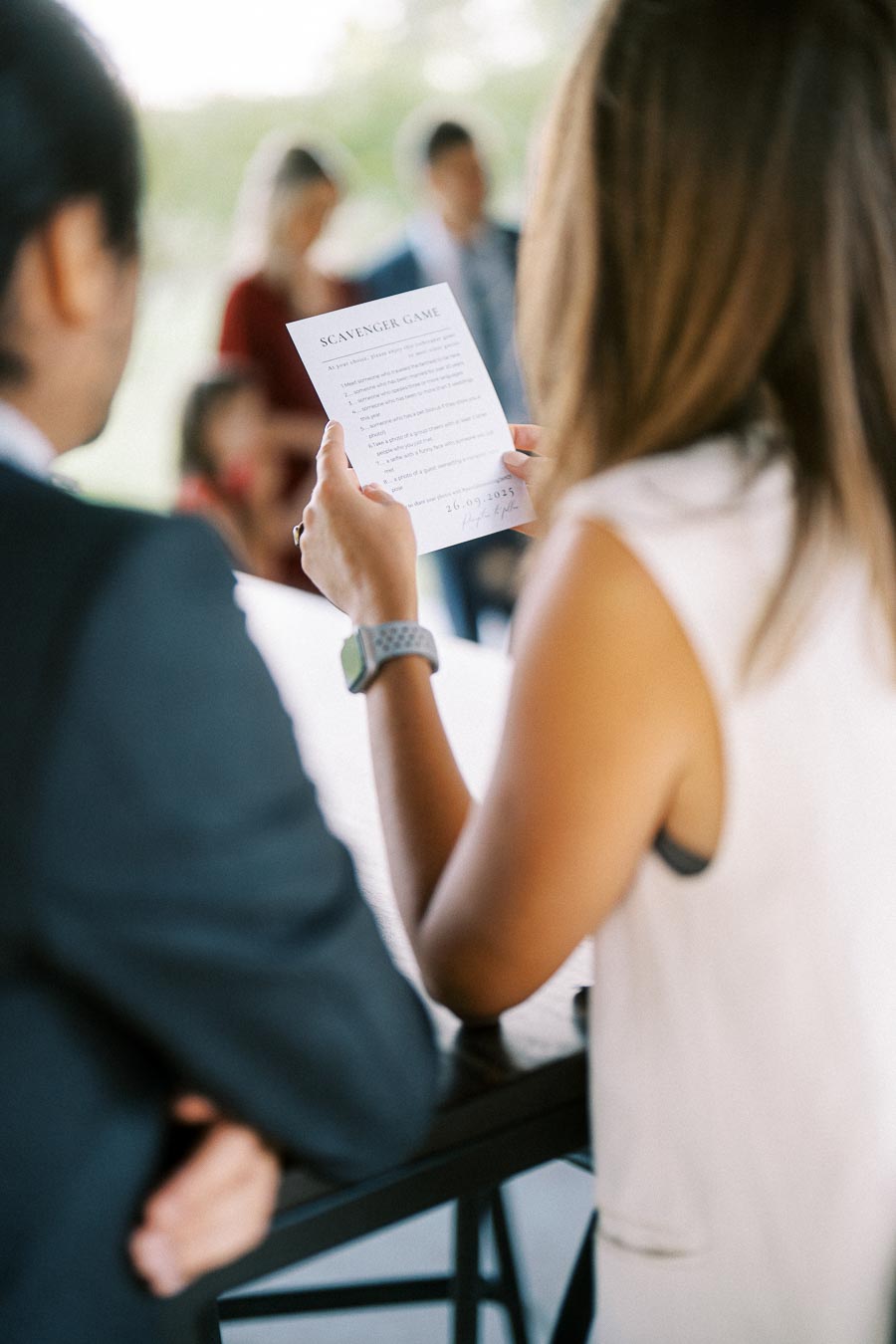 A woman holding a scavenger game paper, with blurred people in the background suggesting a social gathering, possibly an outdoor event on a sunny day.