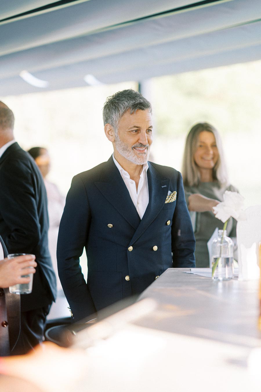 A well-dressed man in a navy suit stands at a stylish event, smiling pleasantly. The soft-focus background shows a woman holding a drink, suggesting a social gathering in a sophisticated setting.