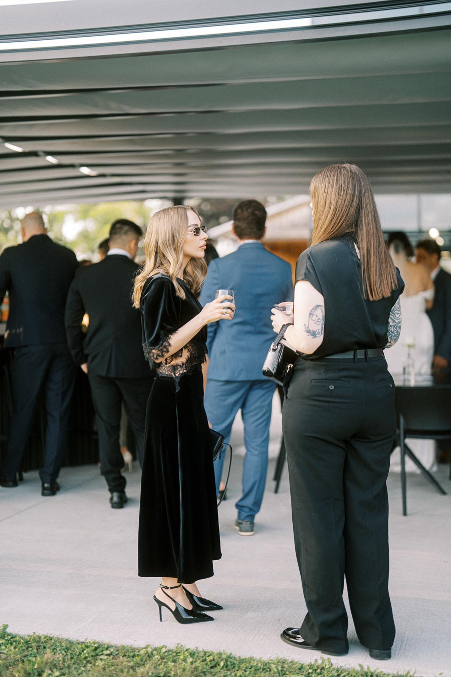 Women in elegant black attire conversing at an outdoor event with other formally dressed attendees showing a classy social gathering atmosphere.
