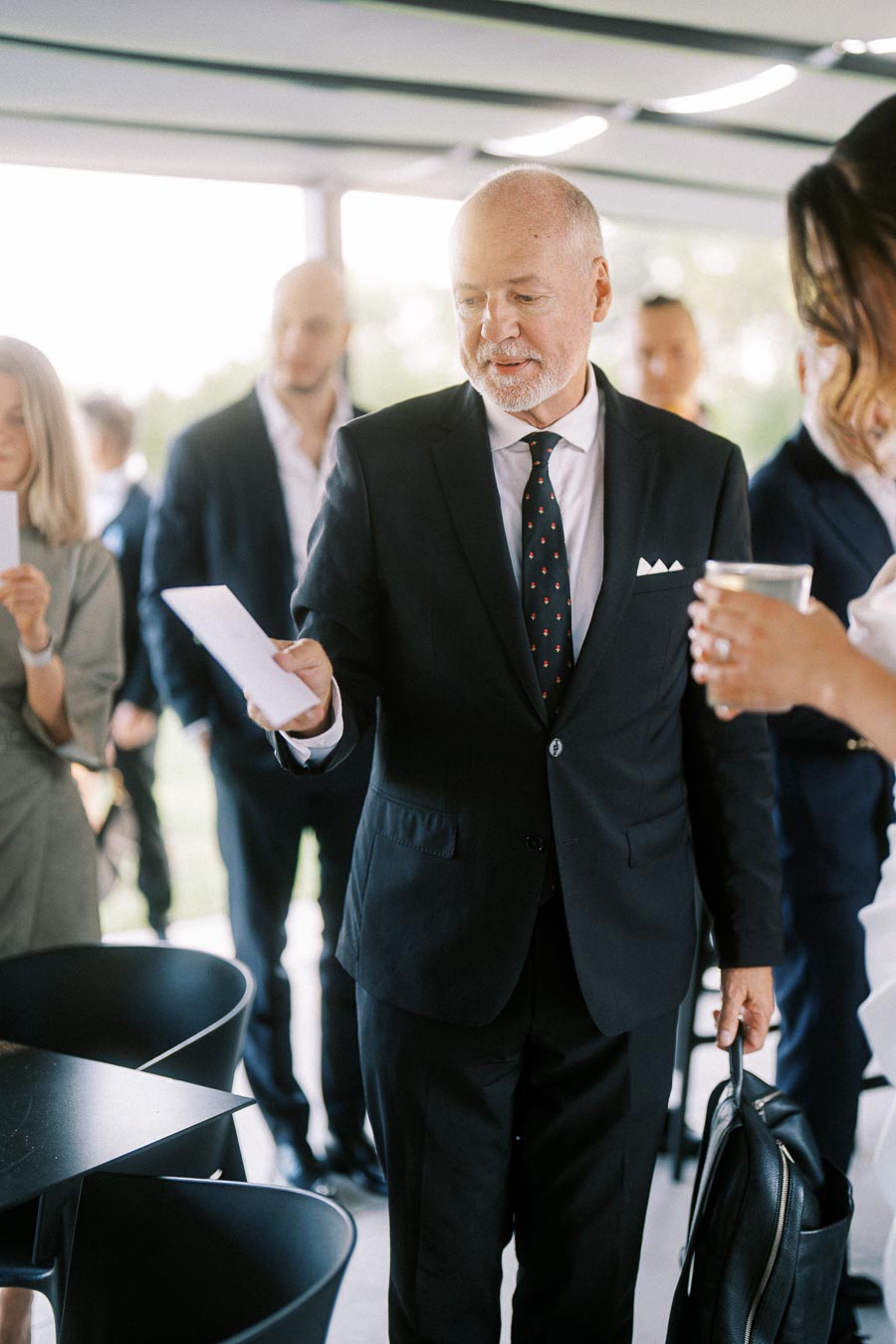 Business professional in a suit holding a piece of paper while interacting with colleagues during a corporate event.