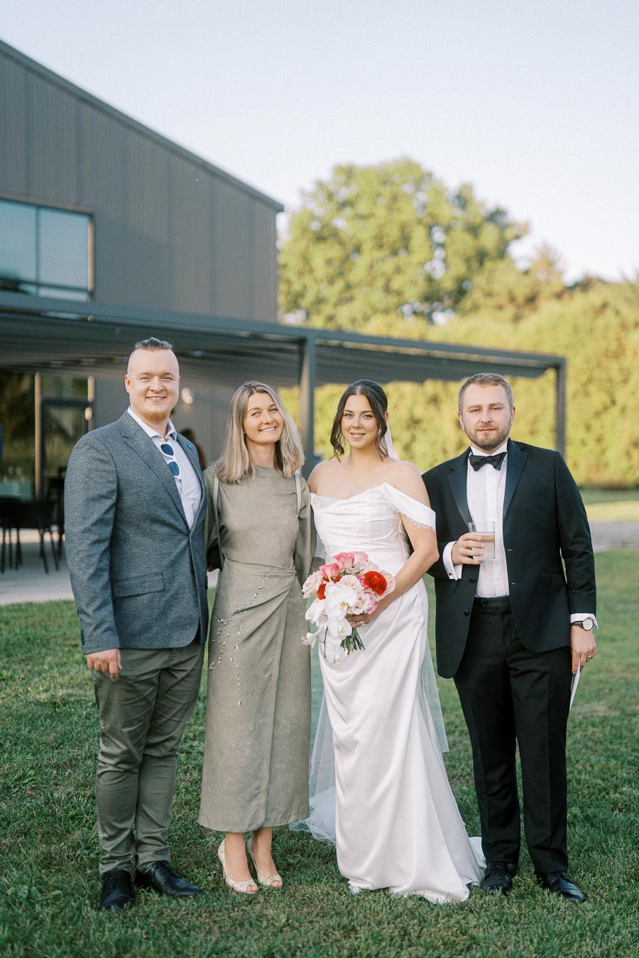 Group photo of a wedding celebration with a bride in a white gown holding a bouquet, standing beside three guests dressed in formal attire, outdoors near a modern building with greenery in the background.